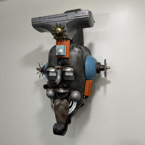 A wall piece constructed of a boat gas tank, aircraft gauges for eyes, vacuum cleaner parts, and a wood chipper part.